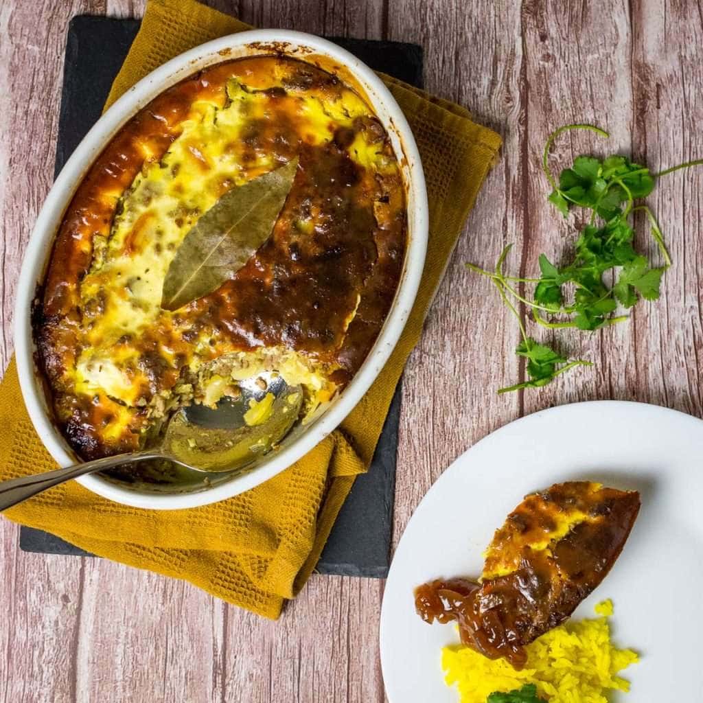 South African Bobotie in a white casserole dish with a portion served on a plate with yellow rice.