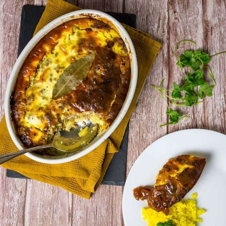 South African Bobotie in a white casserole dish with a portion served on a plate with yellow rice.