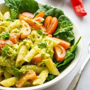Guacamole pasta salad in a white bowl on a bed of spinach.