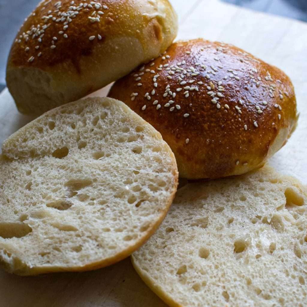 Three sourodugh hamburger buns on a wooden board. One of them is cut in half.