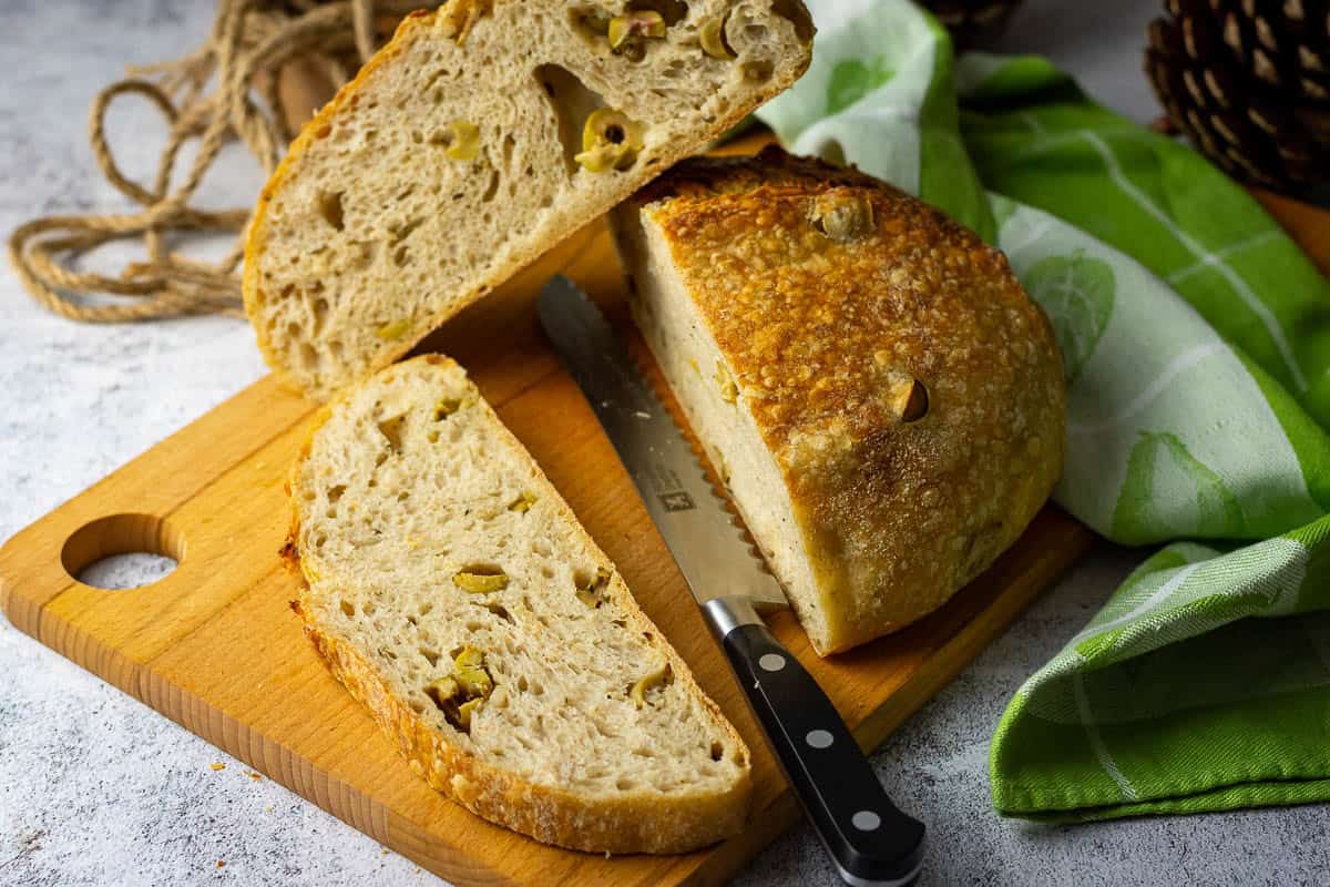 Olive sourdough bread being sliced on a wooden board.