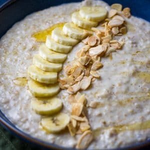Creamy Banana Porridge topped with toasted almonds and banana slices, and drizzled with honey in a dark blue bowl.