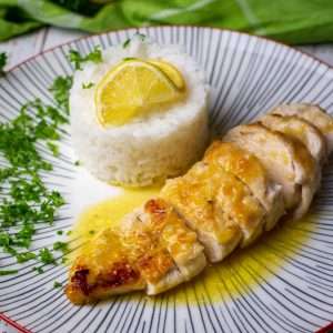 Sliced Chicken Limone with sauce drizzled all over on a white plate with rice, lemon and chopped parsley.