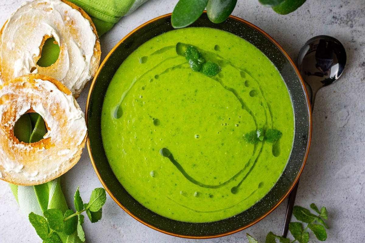 Pea and mint soup in a black bowl with brown rim, with mint oil on top, and a cream cheese bagel next to it.
