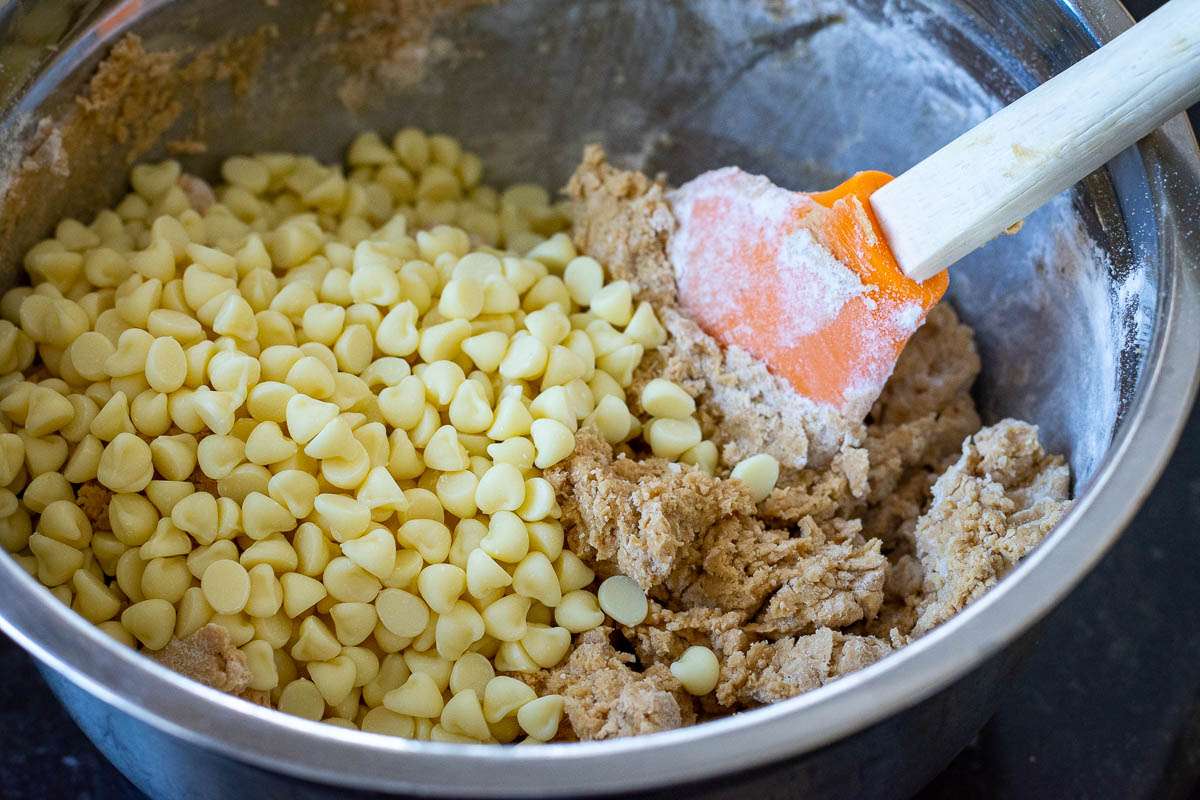 Cookie dough with white chocolate chips on top and an orange rubber spatula in it.