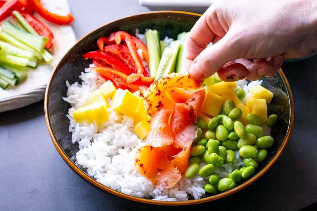 Topping rice and veggies with smoked salmon in a black bowl with brown rim.