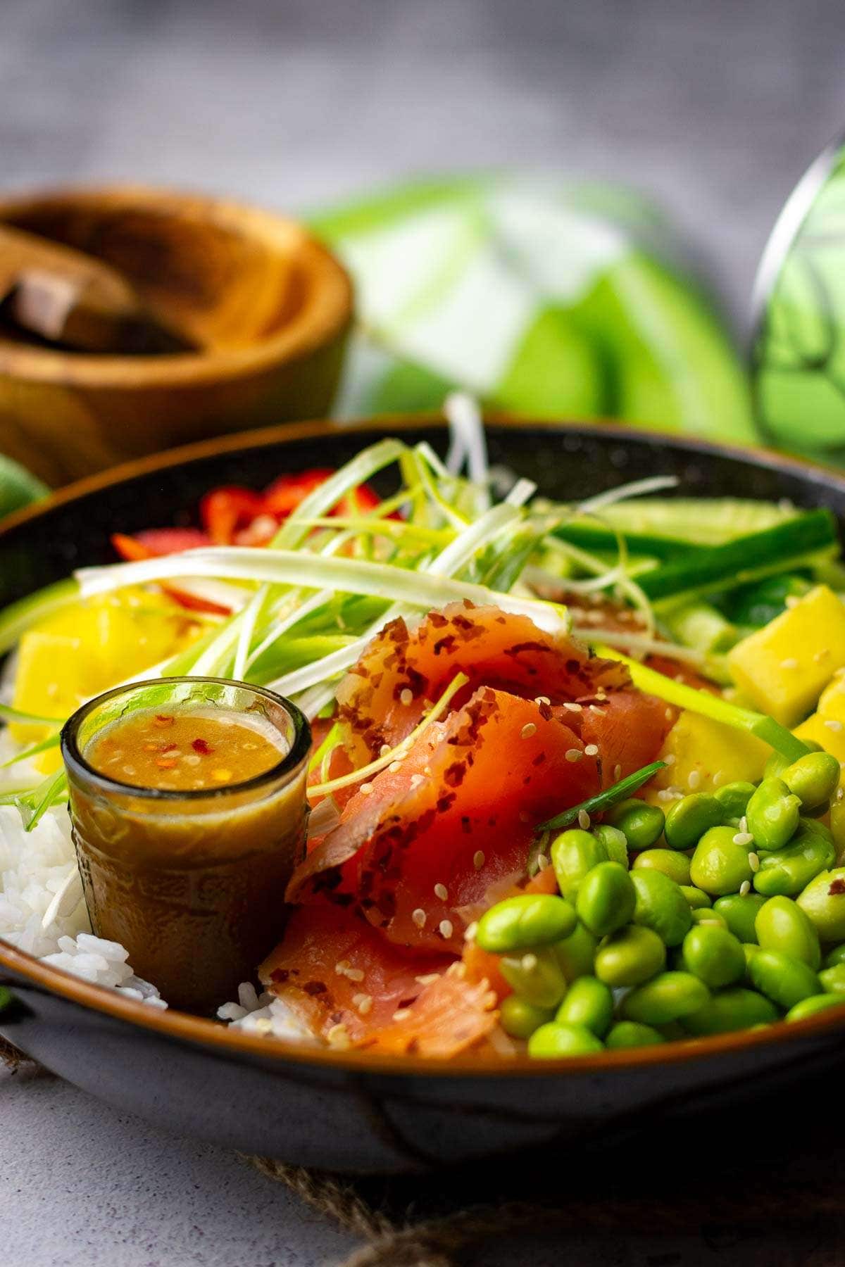 A rice bowl topped with vegetables, mango and smoked salmon with a small shot glass full of dressing.