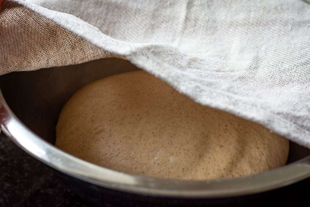 Sourdough Discard Hot Cross Bun dough proofing in a metal bowl under a damp kitchen towel.