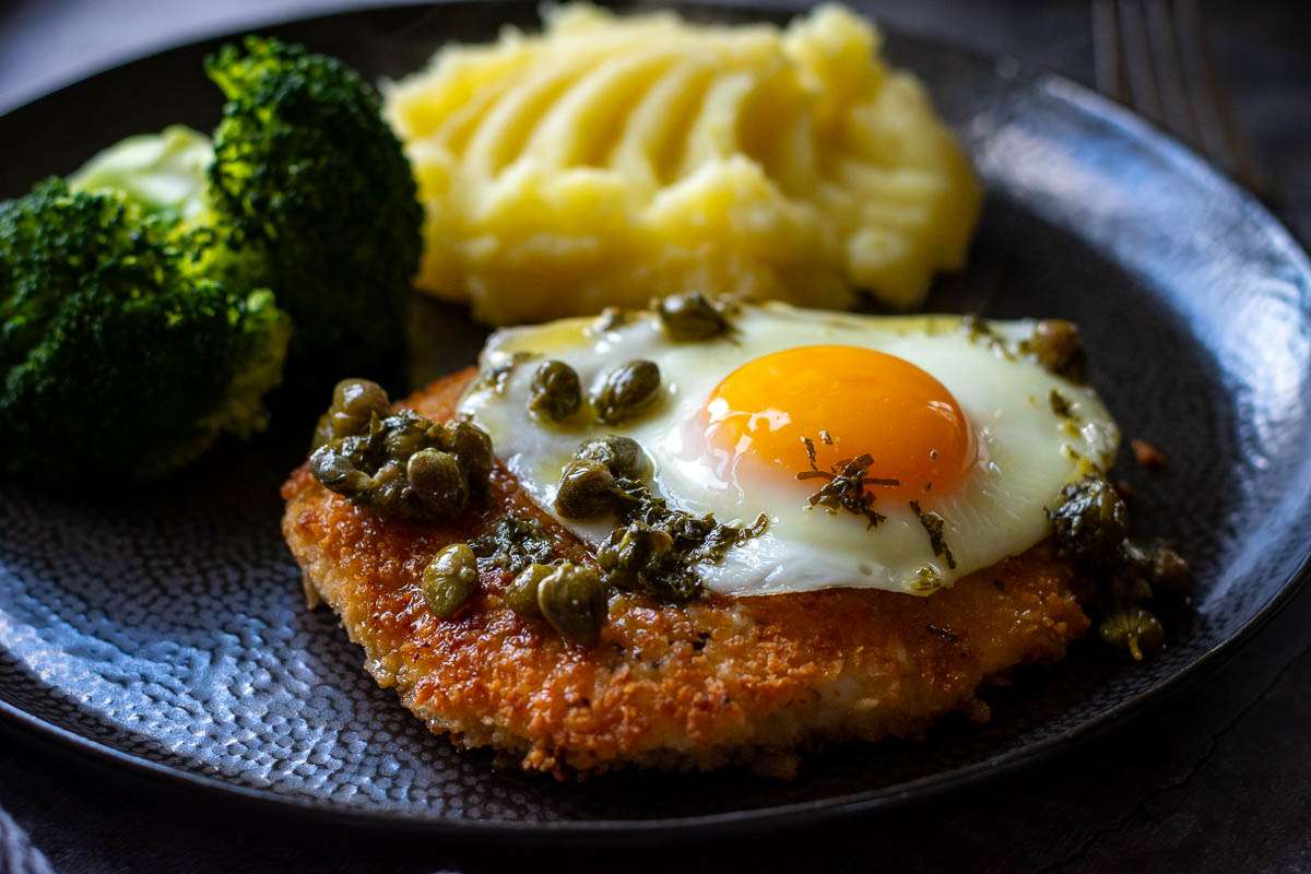 Chicken Schnitzel Holstein with fried egg and caper butter on top served with mashed potatoes on a black plate.