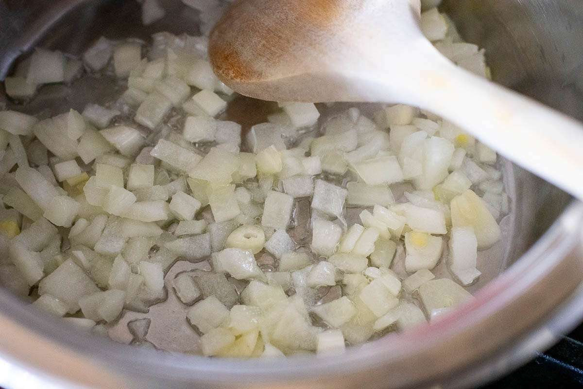 Chopped onions sauteeing in a large pan with a wooden spoon in it.