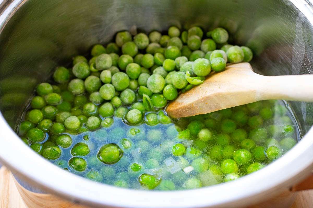 Peas, onions, garlic and stock in a pan with a wooden spoon in it.
