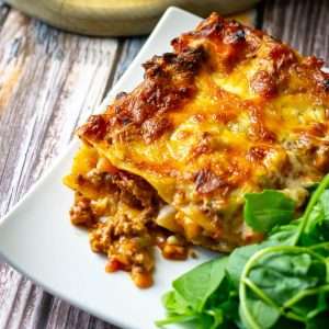A slice of lasagna al forno on a white square place with a green side salad.