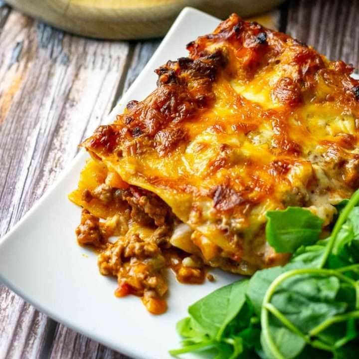 A slice of lasagna al forno on a white square place with a green side salad.
