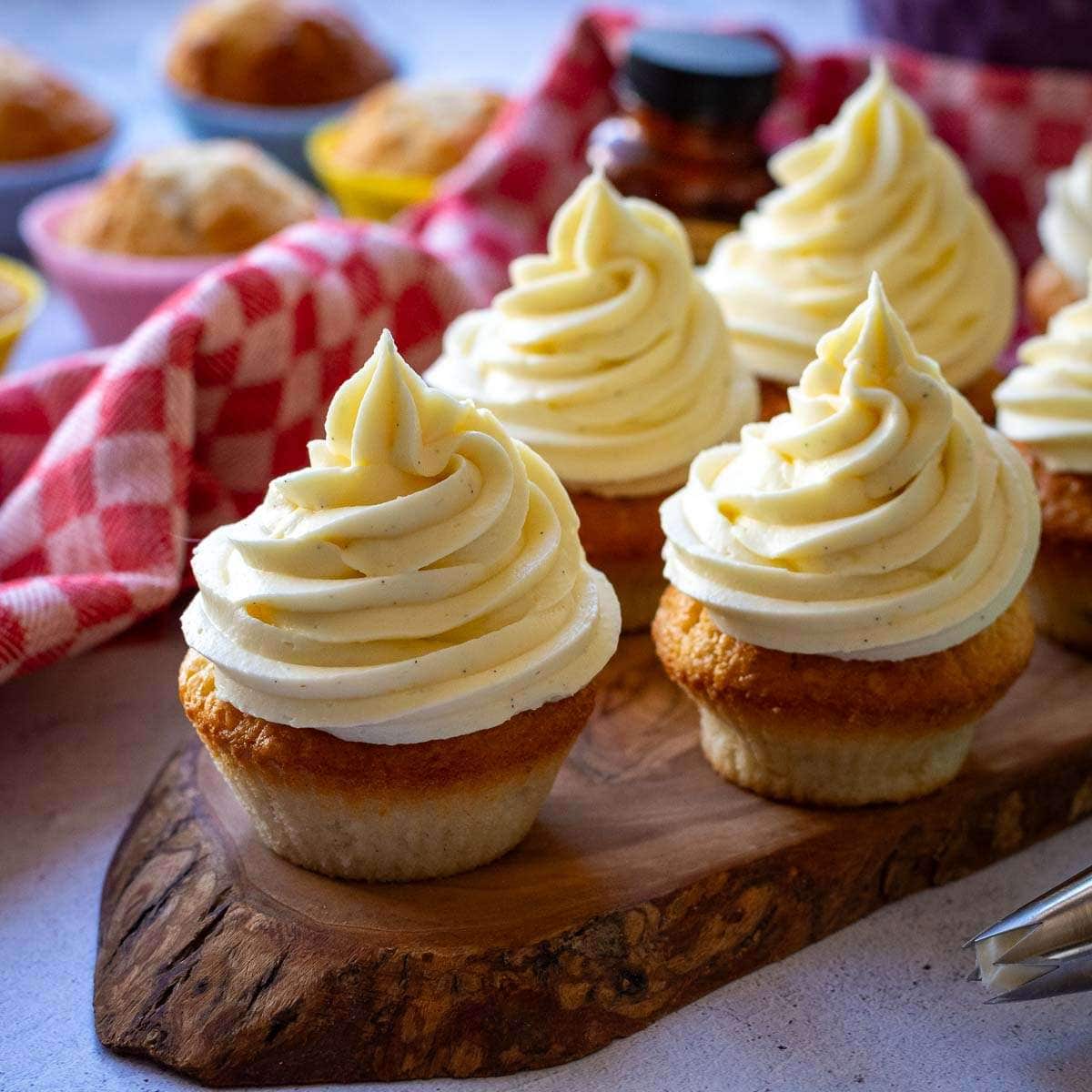 6 sourdough vanilla cupcakes with cream cheese frosting on a wooden board, with plenty more unfrosted cupcakes in the background.