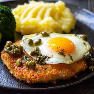 Chicken Schnitzel Holstein with fried egg and caper butter on top served with mashed potatoes on a black plate.