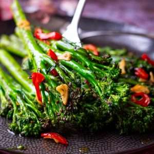 Chargrilled tenderstem broccoli with chilli and garlic being placed on a large black plate.