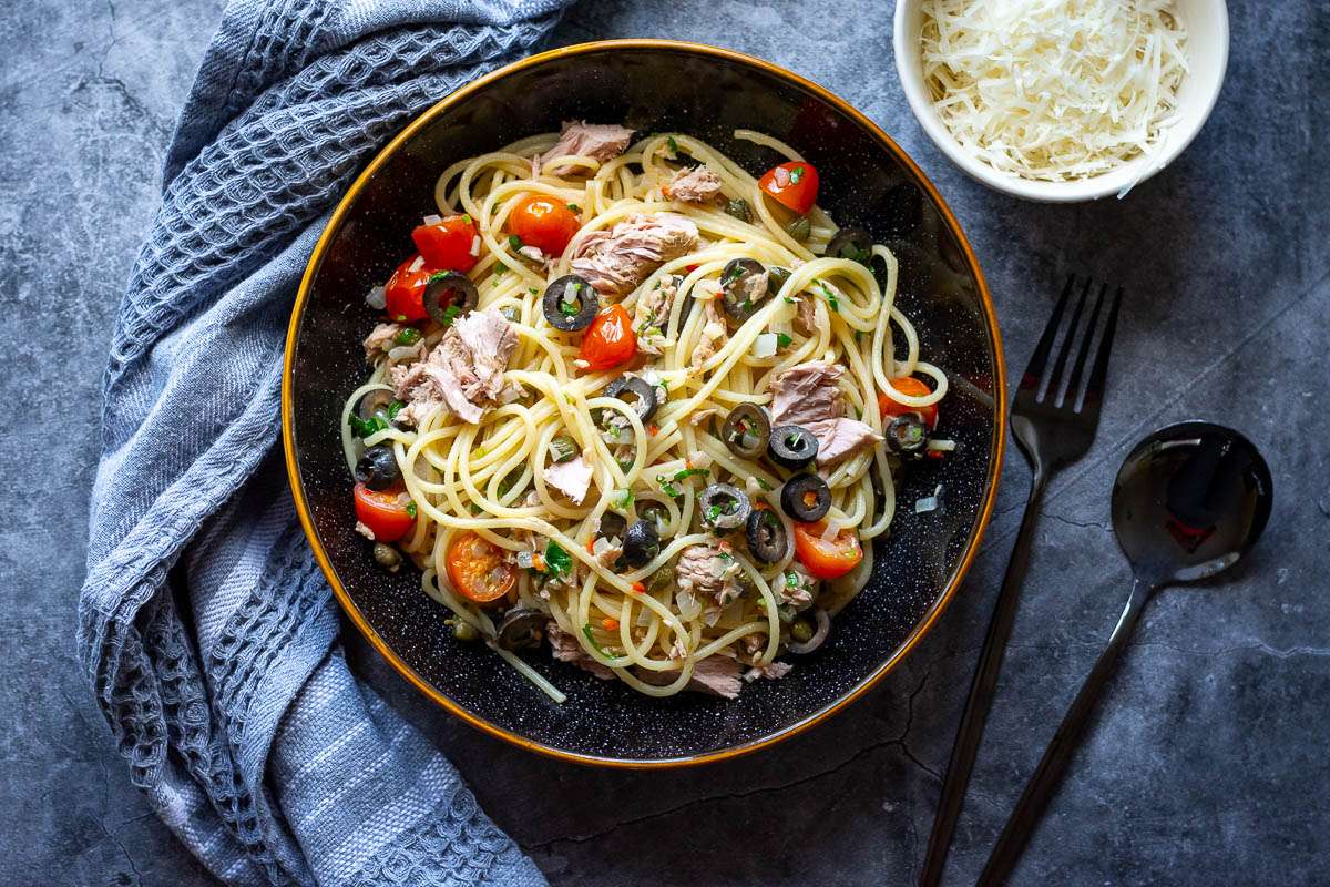 Tuna and Olive pasta with capers and cherry tomatoes in a black bowl with brown rim on a grey surface.