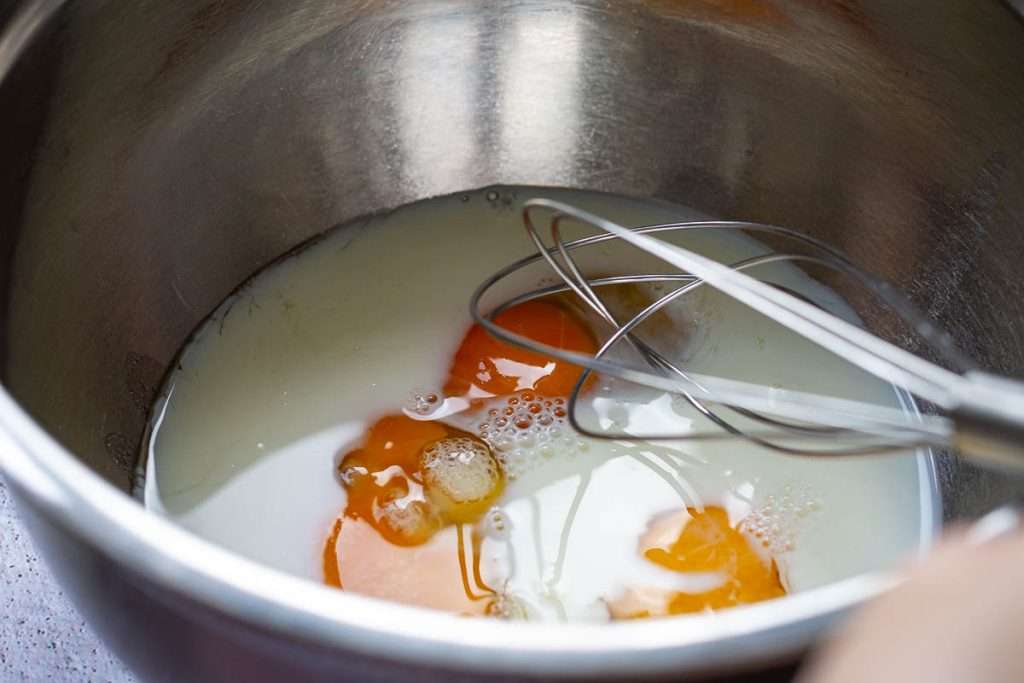 Eggs, sugar and milk being whisked in a large metal bowl with a balloon whisk.
