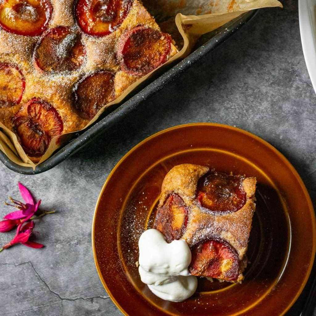 A slice of yogurt plum cake on a small plate with the rest of the cake in a square pan next to it.
