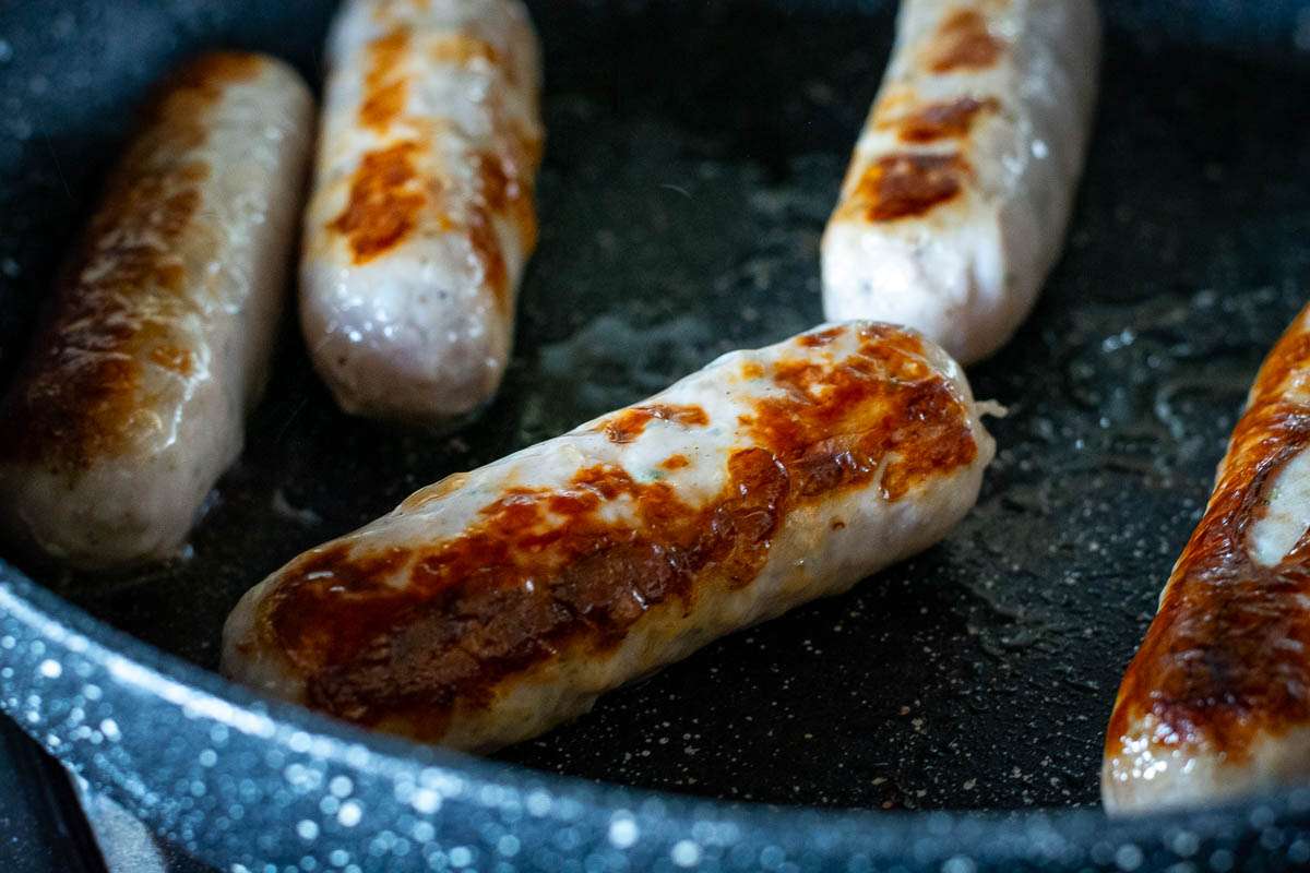 Sausages browning on all sides in a frying pan.