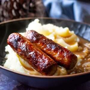 Two sausages with onion gravy all over them and potato mash in a black bowl,