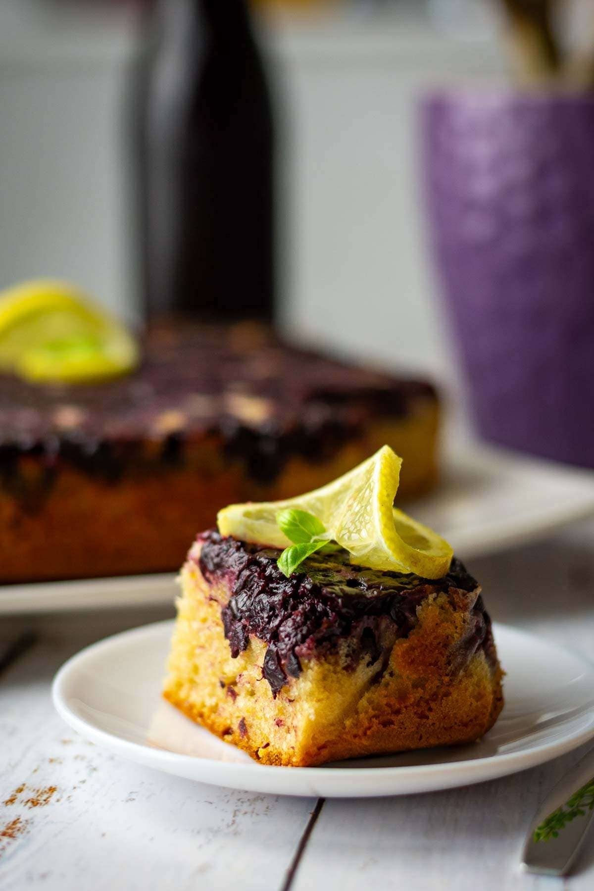 A slice of blackcurrant cake on a white plate with the rest of the cake in the background.