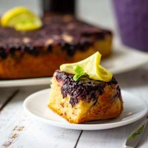 A slice of blackcurrant cake decorated with a lemon slice on a white plate.