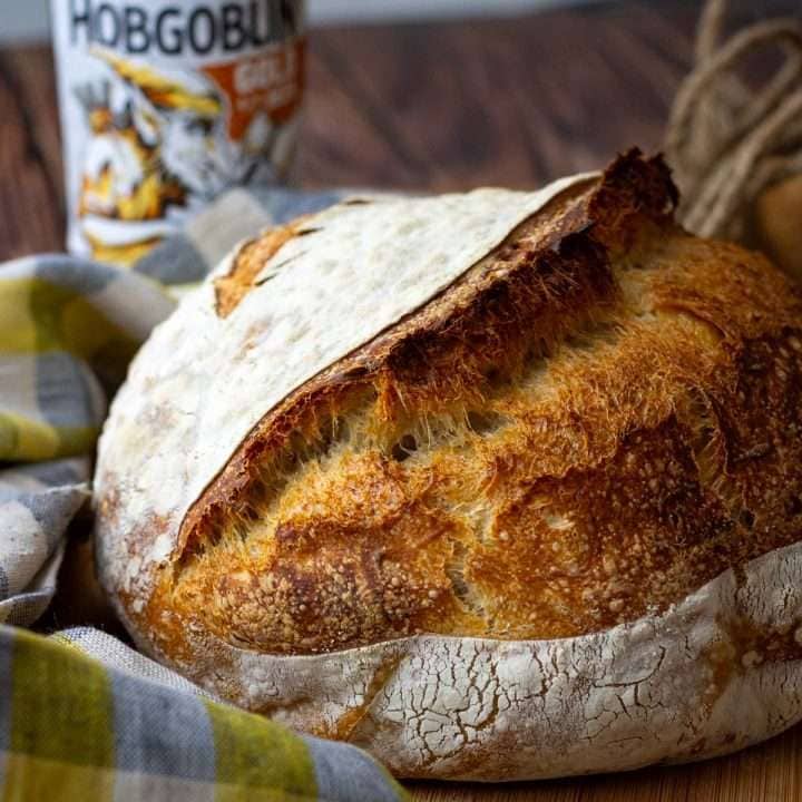 Beer Sourdough Bread with a huge ear on a wooden board next to checked tea towel.