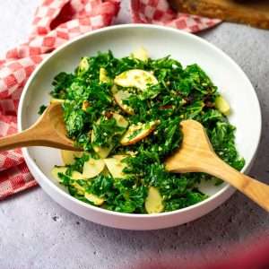 Kale, Apple and Walnut Salad being tossed in a white bowl with wooden serving spoons.