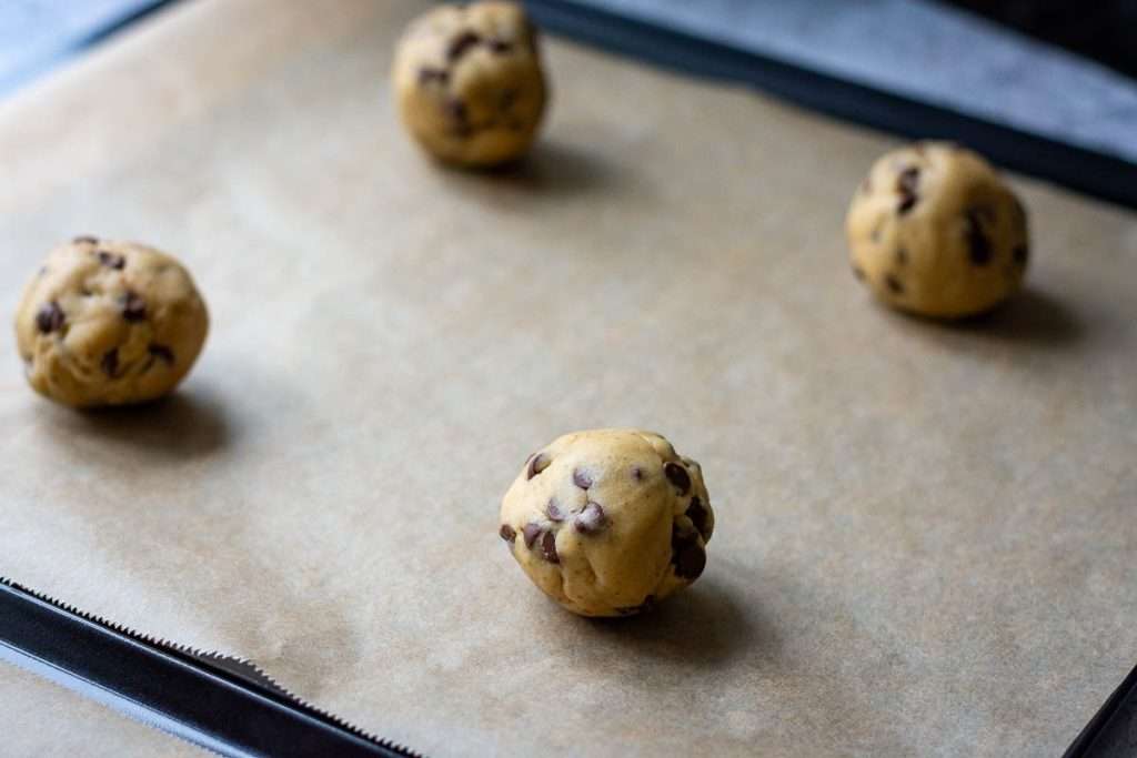 Four chocolate chip cookie dough balls on a parchment lined sheet.