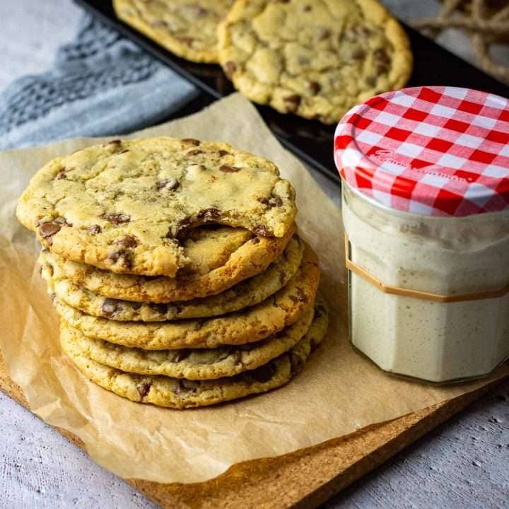 A stack of sourdough discard chocolate chip cookies on a piece of parchment next to a jar of sourdough starter.