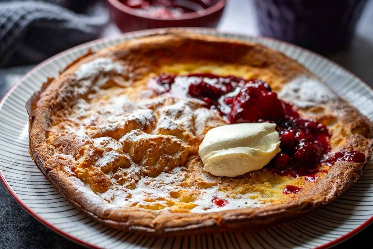 Sourdough Dutch Baby pancake topped with yogurt and berry compote on a white striped plate with dark rim.