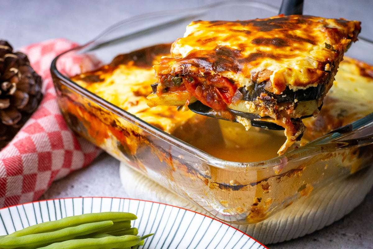 A slice of aubergine bake with cheesy top being lifted out of the ovenproof dish.