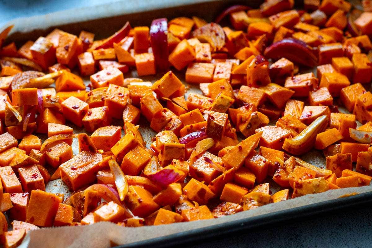 Sweet potatoes and red onions covered in harissa marinade spread on a baking tray.