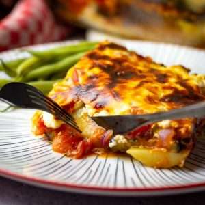 A slice of aubergine and tomato bake with potatoes on a large plate with cutlery approaching.
