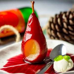 A mulled wine poached pear with one bite missing on a white plate with mulled wine sauce and creme fraiche.