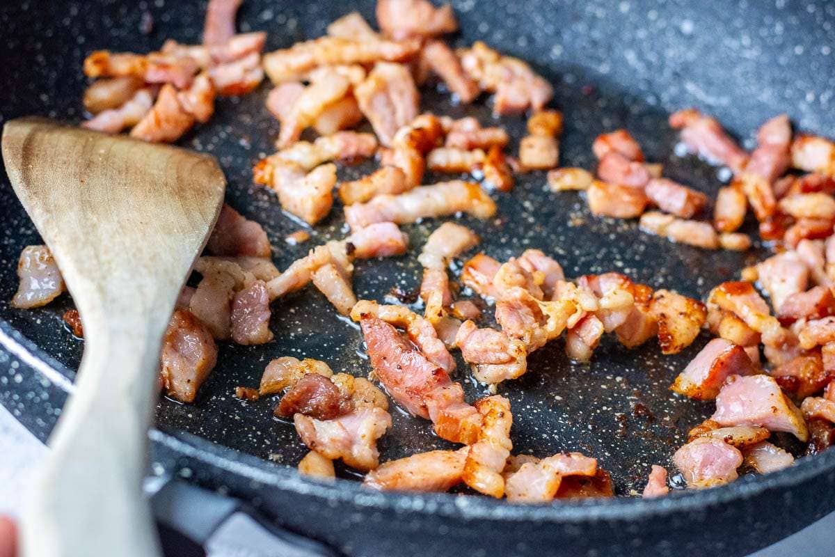 Bacon lardons frying in a large frying pan.