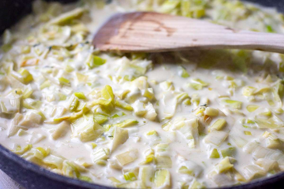 Leeks in creamy sauce in a frying pan with a wooden spatula on top.