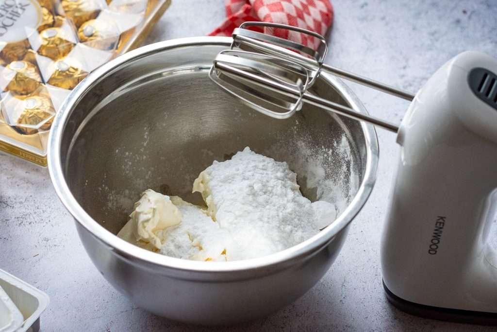 Icing sugar and cream cheese in a metal mixing bowl with a handheld electric mixer next to it.