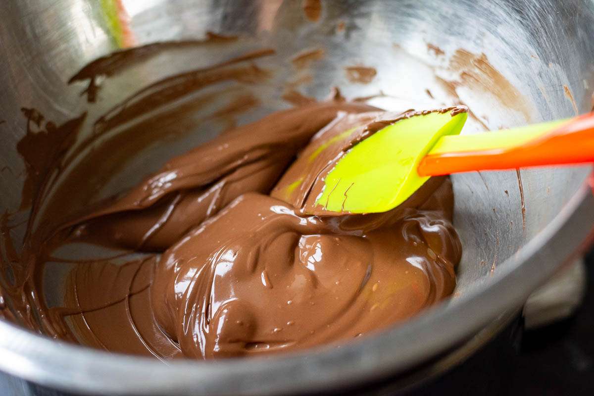Melted milk chocolate in a metal bowl with a green rubber spatula in it.