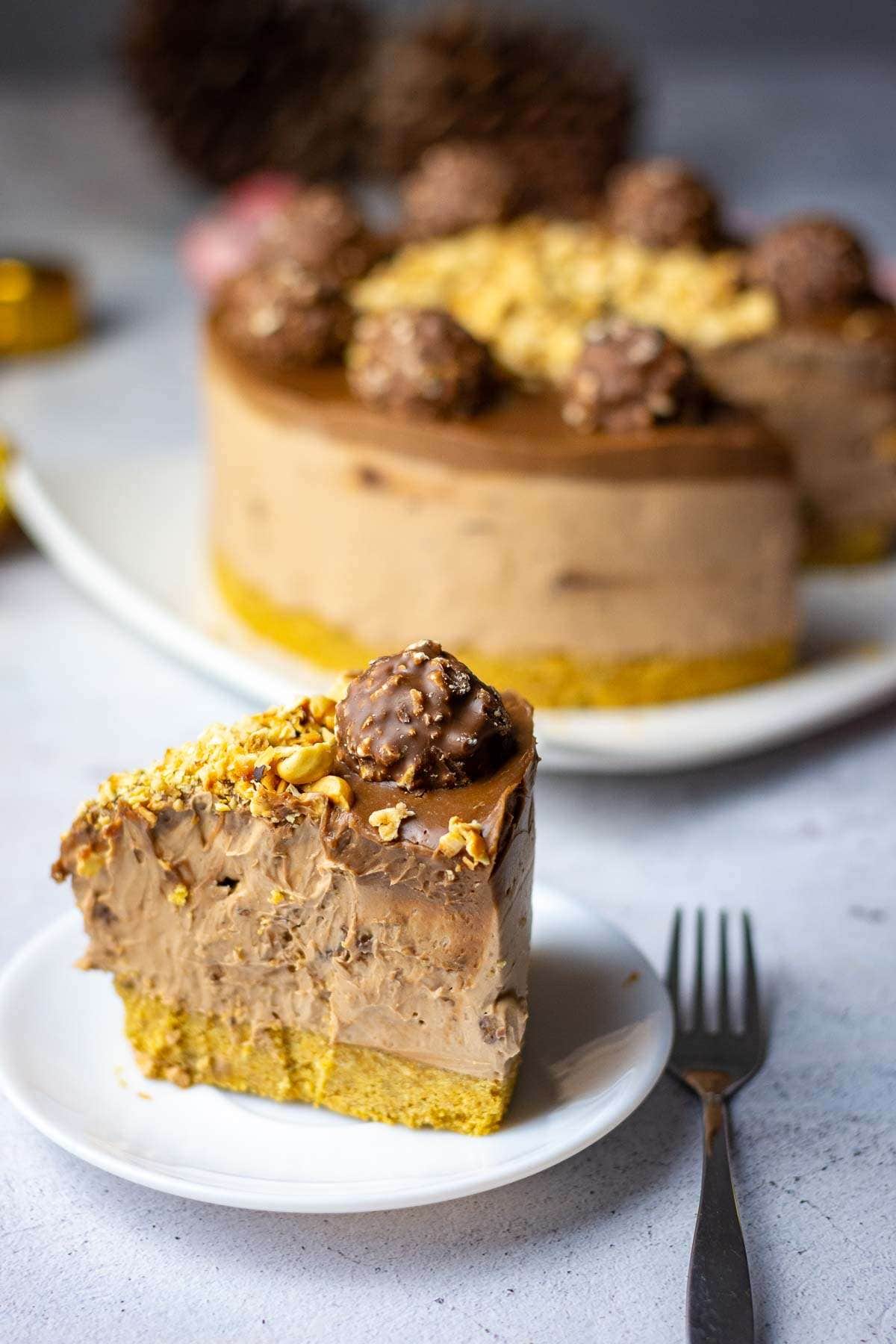 A slice of no-bake Ferrero Rocher Cheesecake on a small white plate with the rest of this no-bake cheesecake in the background.