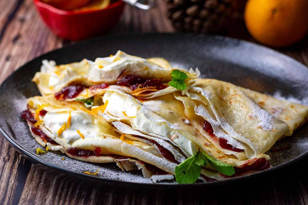Christmas Crepes filled with orange cream and cranberries dusted with icing sugar on a black plate.