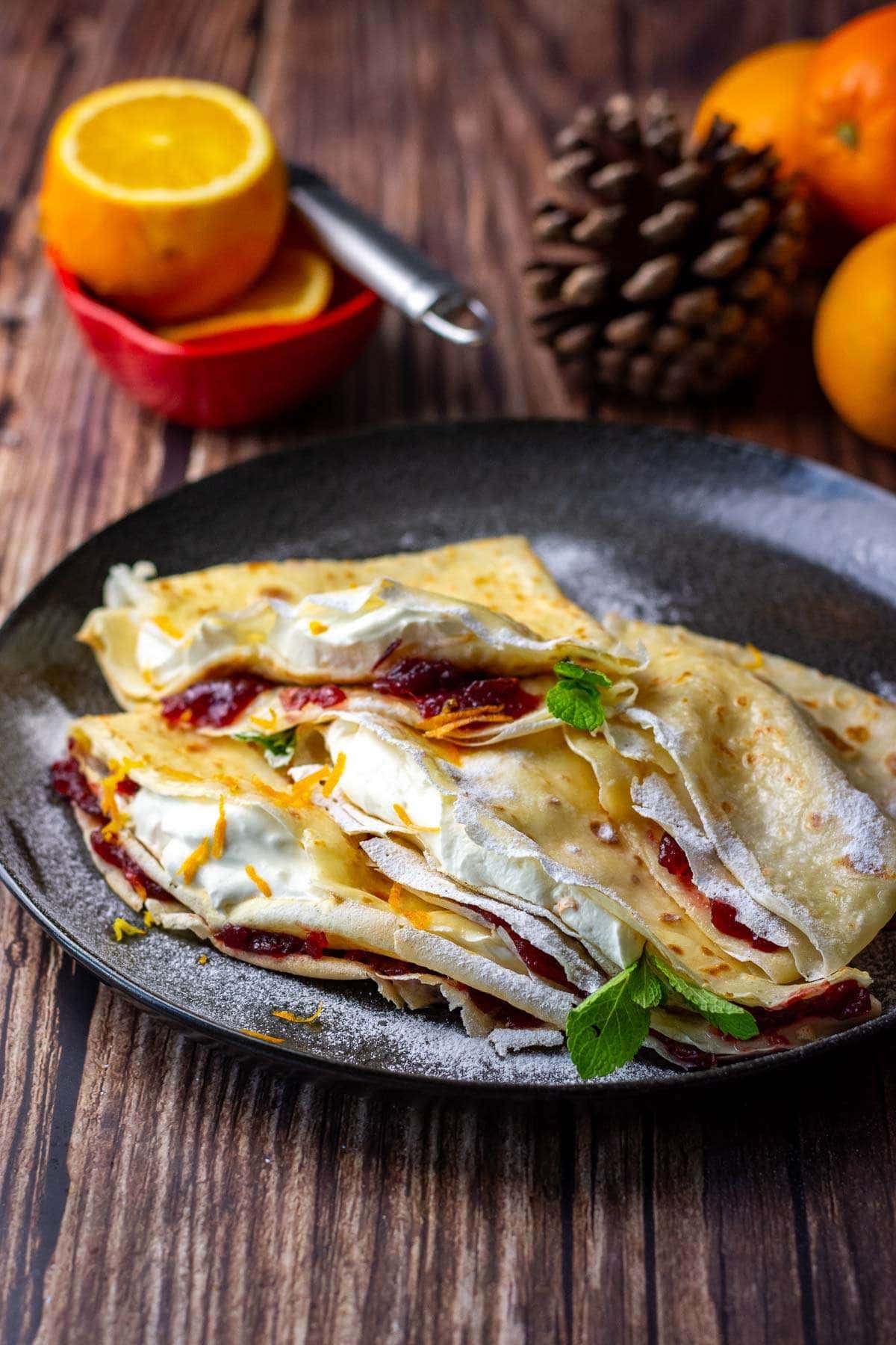Christmas Crepes filled with orange cream and cranberries dusted with icing sugar on a black plate.