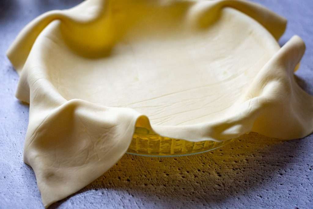 A glass pie dish being lined with shortcrust pastry.