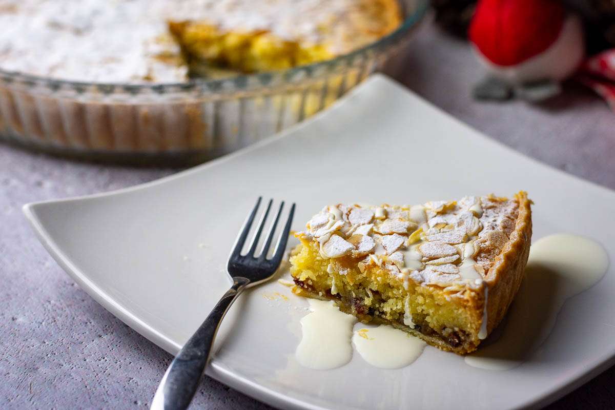 A slice of mincemat frangipane tart being served with a drizzle of cream on a white plate.