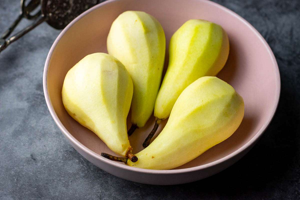 Four peeled pears with stalks intact in a pale pink bowl.
