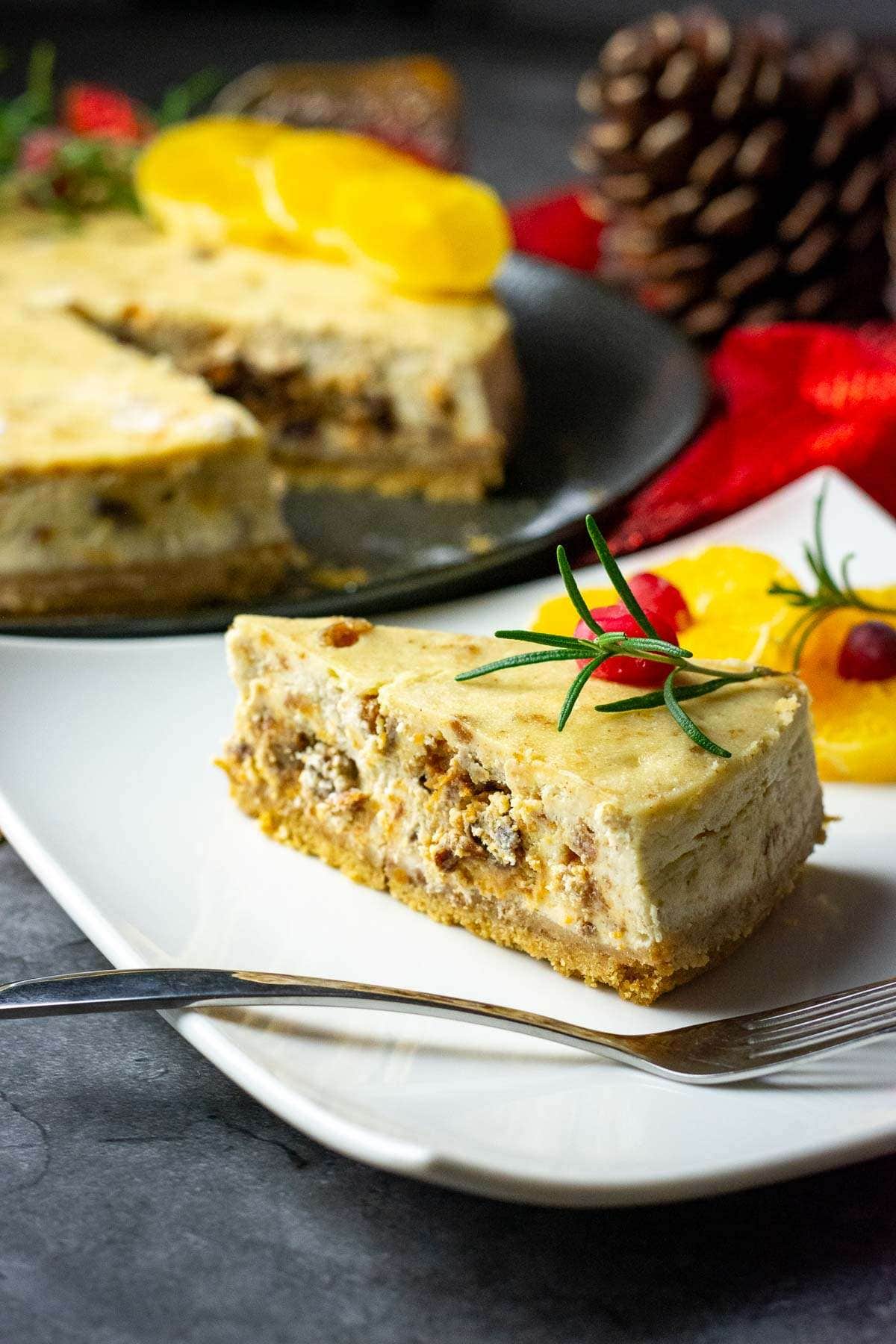 A slice of christmas cake cheesecake on a white square plate garnished with rosemary and cranberries.