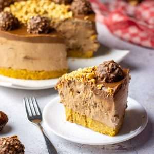 A slice of no-bake Ferrero Rocher Cheesecake on a small white plate with the rest of this no-bake cheesecake in the background.