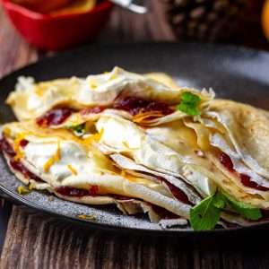 Christmas Crepes filled with orange cream and cranberries dusted with icing sugar on a black plate.