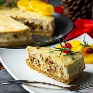 A slice of christmas cake cheesecake on a white square plate garnished with rosemary and cranberries.